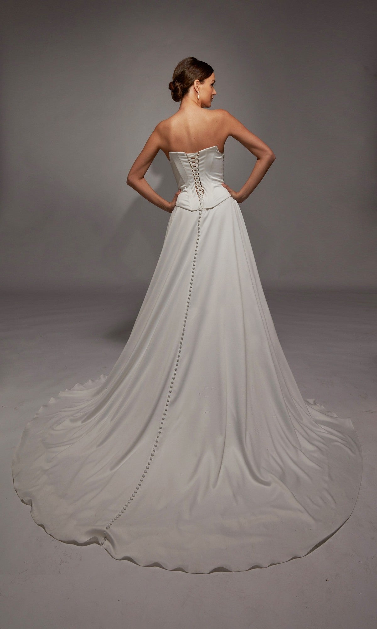 A woman faces away, wearing the Long White Dress: Alyce 7142— a satin bridal gown featuring a fitted corset bodice, lace-up back, and a long train with buttons down the center.